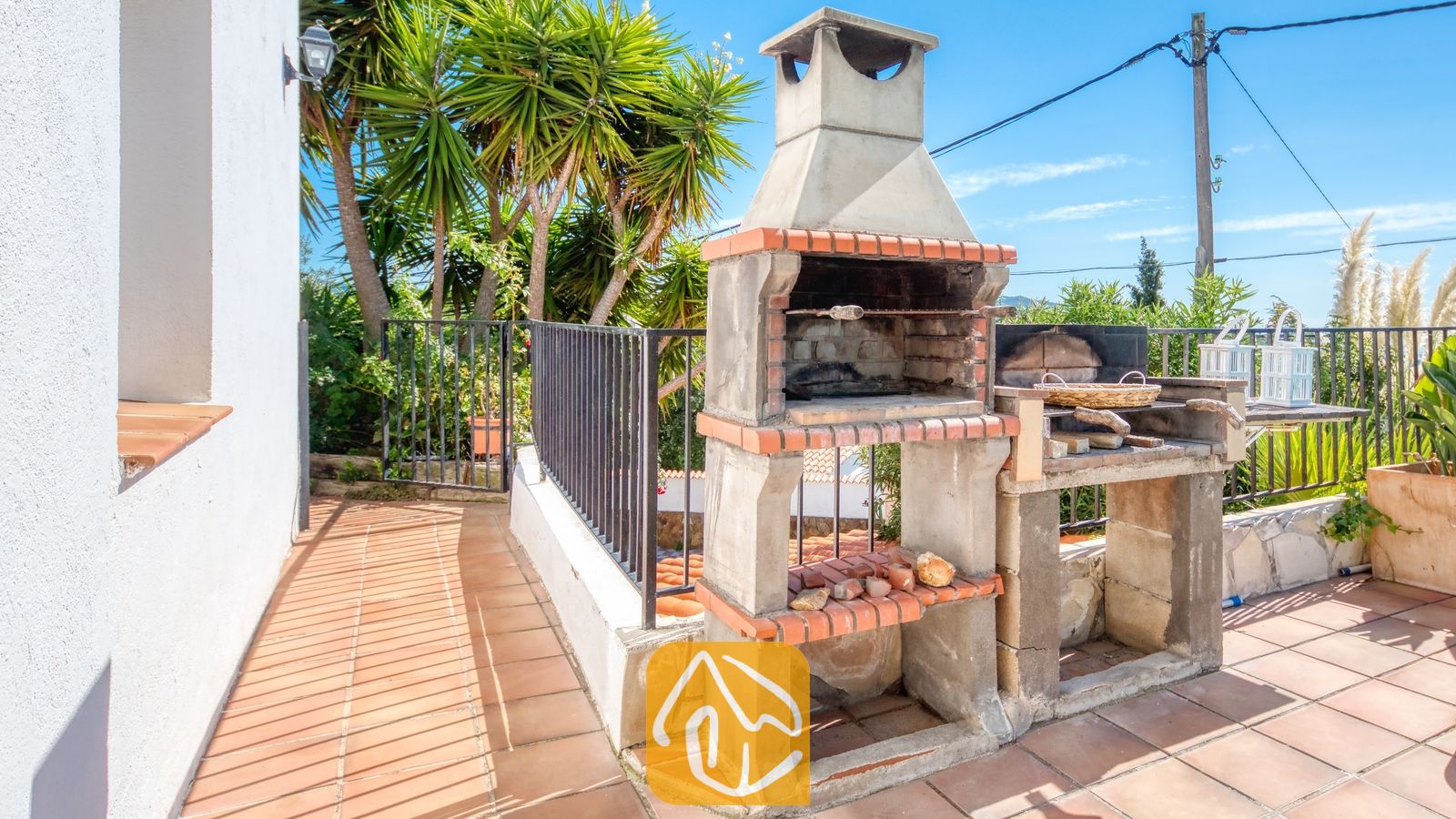 Vakantievilla's Costa Brava Spanje - Villa Sofia - Buiten stenen barbecue met een bakstenen schoorsteen op een betegeld terras bij Villa Sofia, met palmbomen op de achtergrond.