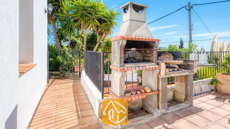 Vakantievilla's Costa Brava Spanje - Villa Sofia - Buiten stenen barbecue met een bakstenen schoorsteen op een betegeld terras bij Villa Sofia, met palmbomen op de achtergrond.