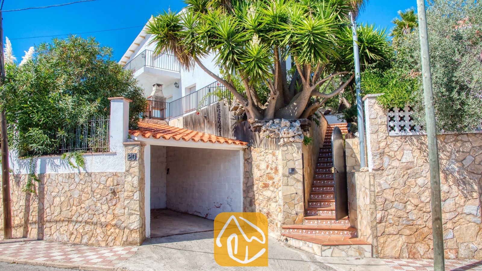 Vakantievilla's Costa Brava Spanje - Villa Sofia - Buitenkant van Villa Sofia met een overdekte garage, stenen muur, betegelde buitentrap en een grote palmboom tegen een blauwe lucht.