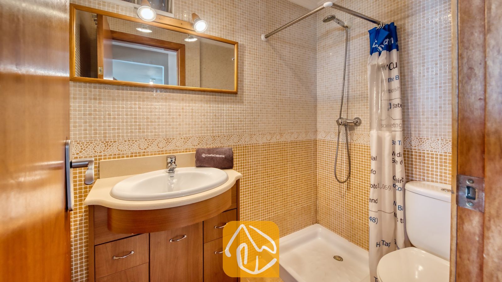 Vakantievilla's Costa Brava Spanje - Villa Sofia - Badkamer voorzien van kleine mozaïektegels, een houten badkamermeubel met een witte wastafel, een spiegel met twee wandlampen en een inloopdouche met een douchegordijn.