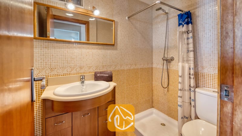 Villas de vacances Costa Brava Espagne - Villa Sofia - Salle de bains habillée de petits carreaux de mosaïque, dotée d'un meuble sous-vasque en bois avec un lavabo blanc, d'un miroir encadré de deux appliques murales et d'une douche à l'italienne avec rideau.