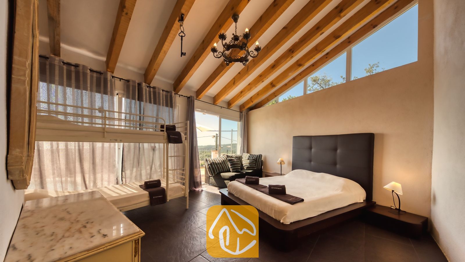 Vakantievilla's Costa Brava Spanje - Villa Sofia - Ruime slaapkamer in Villa Sofia met authentieke houten plafondbalken, een wit metalen stapelbed en een groot tweepersoonsbed met een donker leren hoofdeinde.
