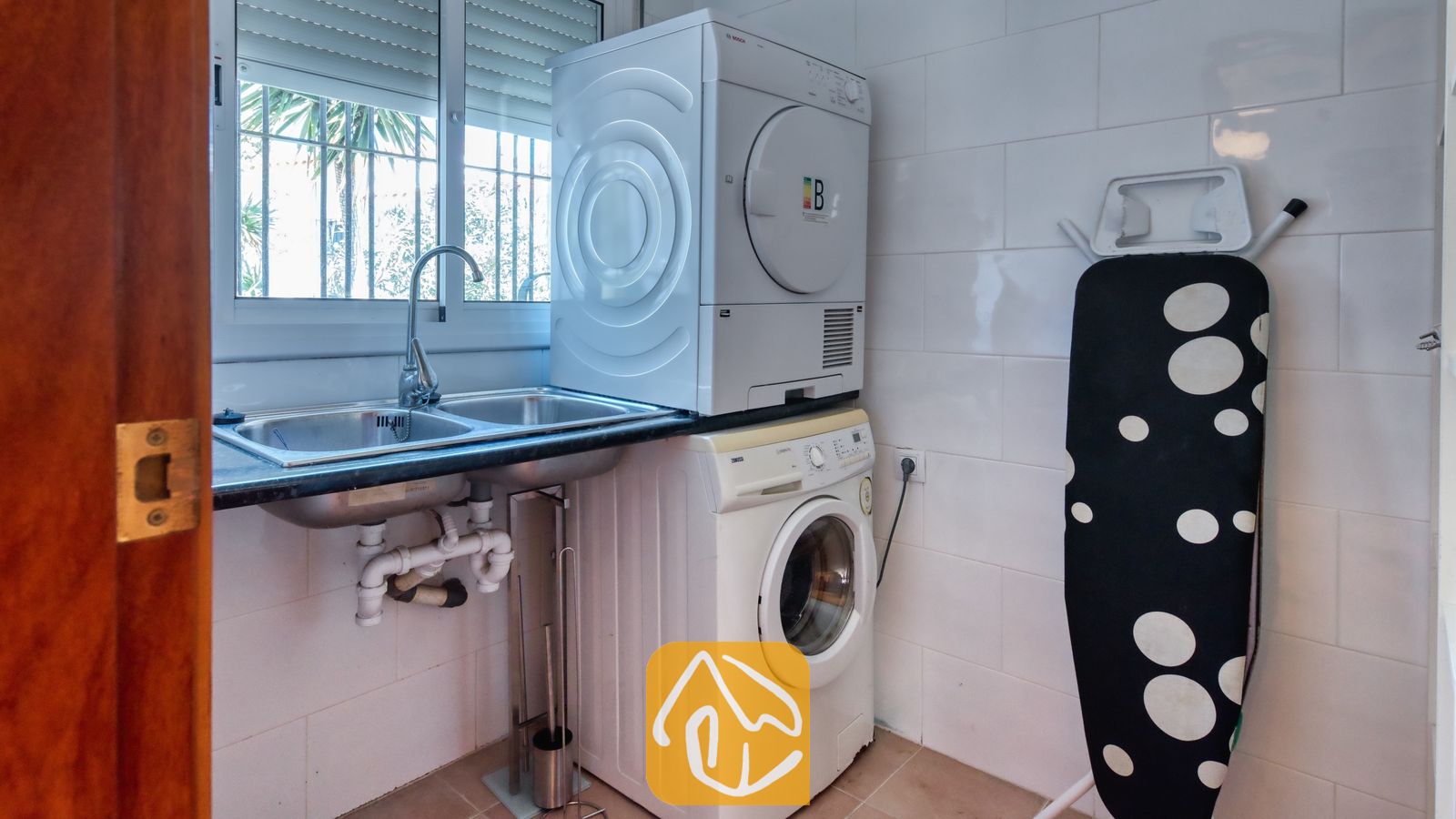 Vakantievilla's Costa Brava Spanje - Villa Sofia - Bijkeuken met een gestapelde wasdroogcombinatie, dubbele spoelbak, witte tegelmuren en een inklapbare strijkplank.