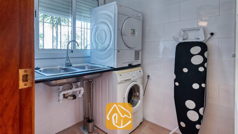 Villas de vacances Costa Brava Espagne - Villa Sofia - Buanderie équipée d'un lave-linge et d'un sèche-linge superposés, d'un évier à double bac, de murs carrelés de blanc et d'une table à repasser murale repliable.