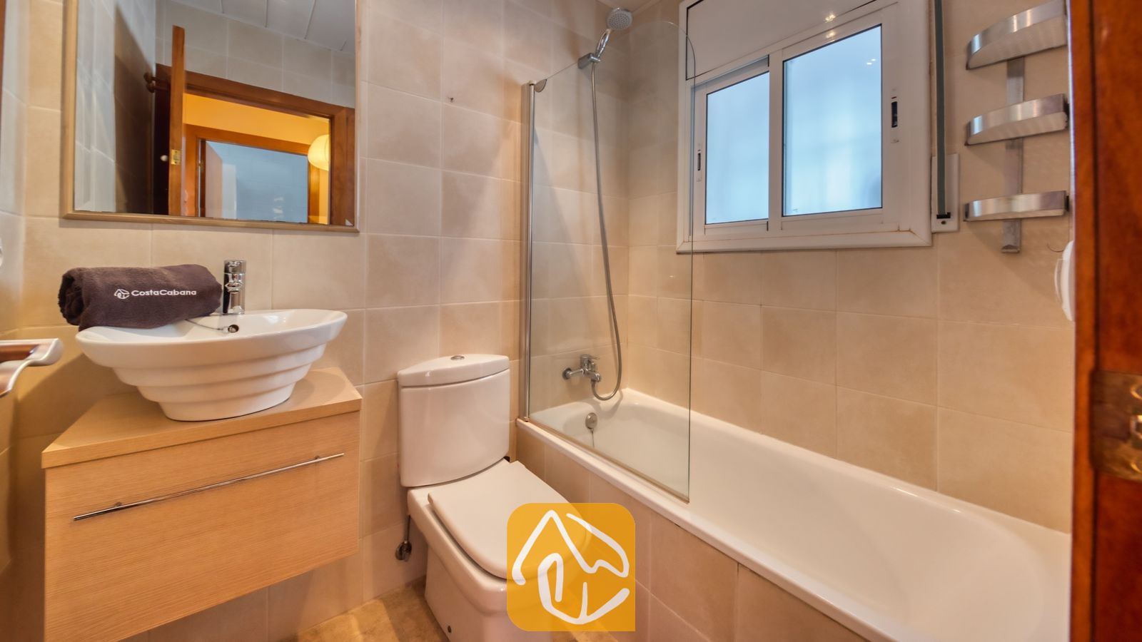 Vakantievilla's Costa Brava Spanje - Villa Sofia - Betegelde badkamer voorzien van een wit ligbad, een glazen douchewand, een toilet en een modern badkamermeubel met een grijze handdoek.