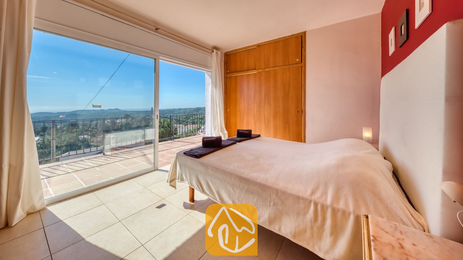 Vakantievilla's Costa Brava Spanje - Villa Sofia - Slaapkamer in Villa Sofia met een tweepuntssbed voorzien van opgerolde handdoeken, een houten kledingkast en glazen schuifdeuren die opengaan naar een betegeld balkon met uitzicht op de verre heuvels.