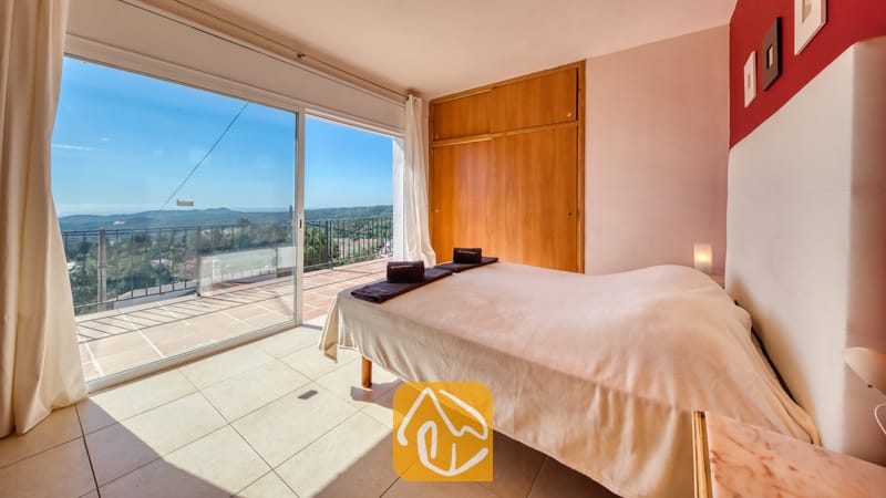 Villas de vacances Costa Brava Espagne - Villa Sofia - Chambre de la Villa Sofia comprenant un lit double avec des serviettes roulées, une armoire en bois et des portes coulissantes en verre s'ouvrant sur un balcon carrelé surplombant des collines lointaines.