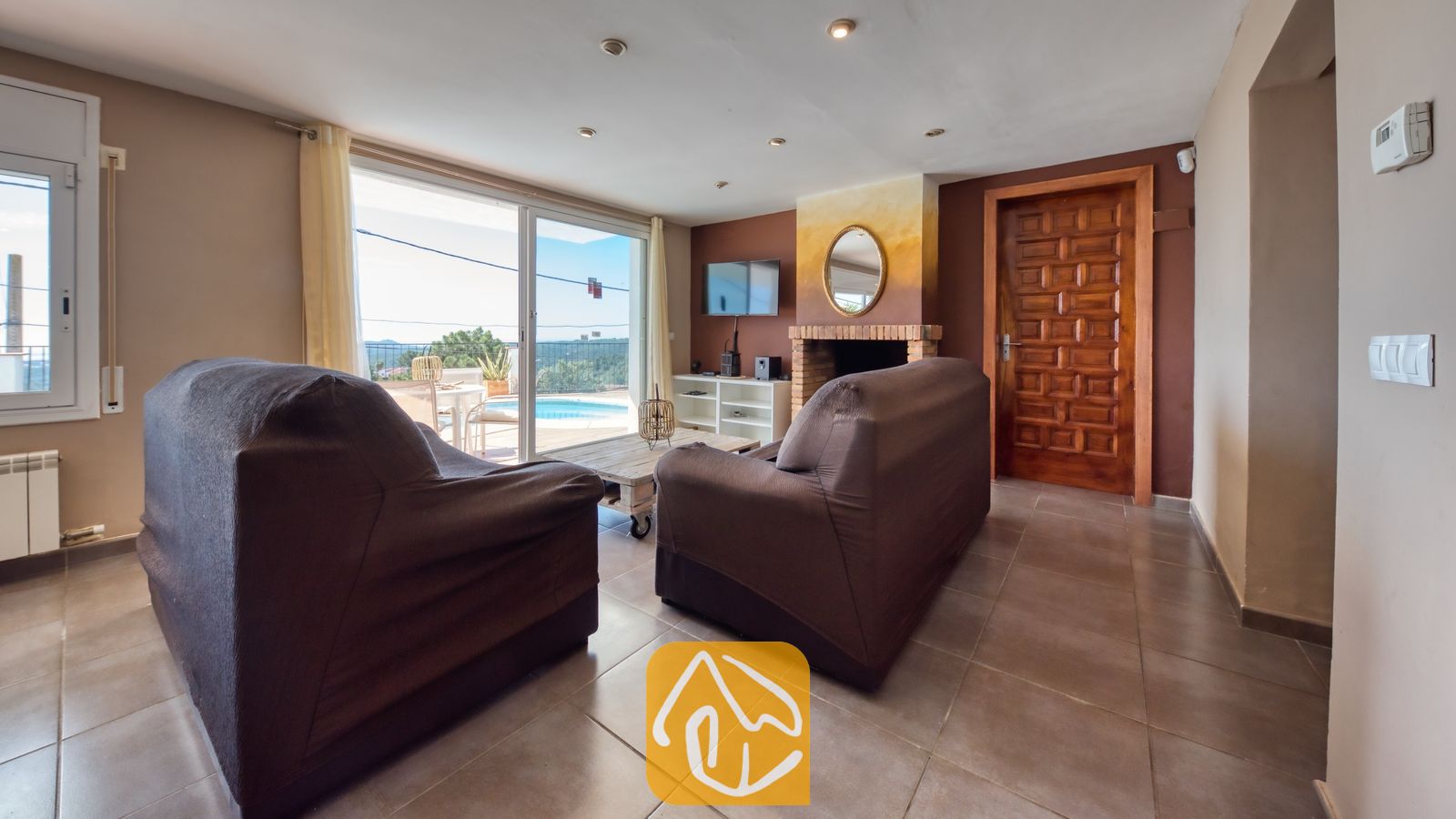 Vakantievilla's Costa Brava Spanje - Villa Sofia - Woonkamer van Villa Sofia met twee bruine gestoffeerde banken, een houten salontafel, een open haard van baksteen en glazen schuifdeuren die opengaan naar een zwembadterras.