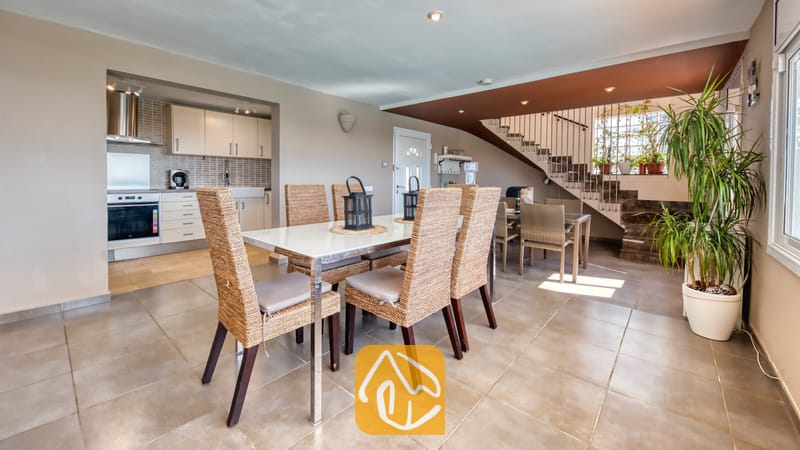 Vakantievilla's Costa Brava Spanje - Villa Sofia - Eethoek met een glazen tafel en gevlochten rotan stoelen bij Villa Sofia, voorzien van tegelvloeren en een open keuken op de achtergrond.