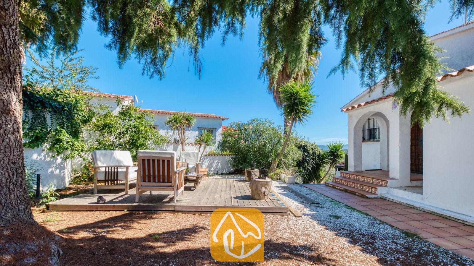 Vakantievilla's Costa Brava Spanje - Villa Sofia - Houten terras met fauteuils in de zonnige tuin van Villa Sofia, voorzien van witte buitenmuren, betegelde treden van de veranda en dennenbomen.