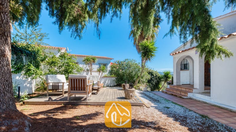 Vakantievilla's Costa Brava Spanje - Villa Sofia - Houten terras met fauteuils in de zonnige tuin van Villa Sofia, voorzien van witte buitenmuren, betegelde treden van de veranda en dennenbomen.