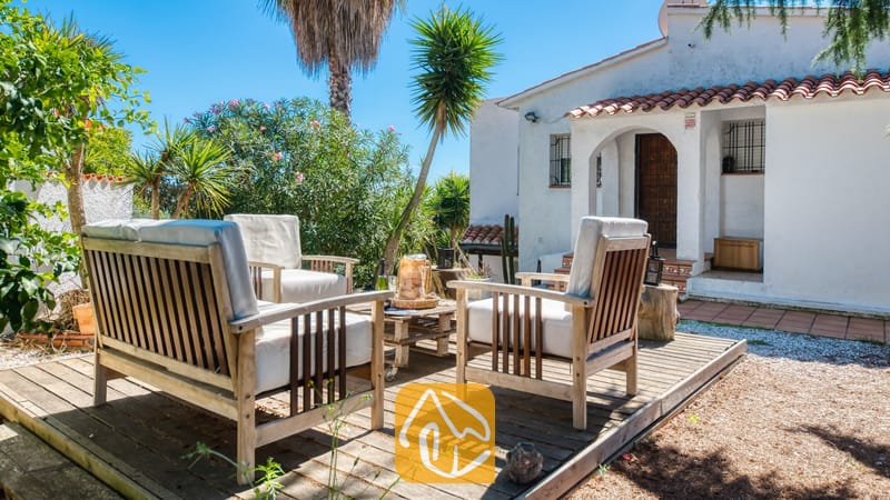 Vakantievilla's Costa Brava Spanje - Villa Sofia - Houten terrasmeubilair met witte kussens op een houten vlonder voor Villa Sofia, met een witte buitenmuur, terracotta dakpannen en palmbomen onder een helderblauwe hemel.