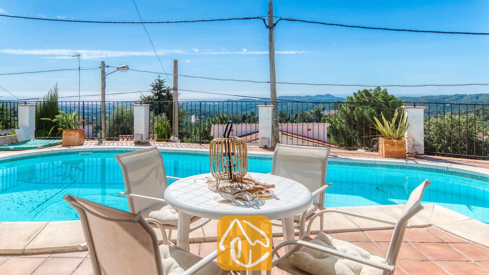 Vakantievilla's Costa Brava Spanje - Villa Sofia - Buitenw Eetgedeelte bij het zwembad van Villa Sofia, voorzien van een witte kunststof tafel, vier bijpassende stoelen met kussens en een rieten lantaarn als middelpunt, met uitzicht op de verre heuvels.