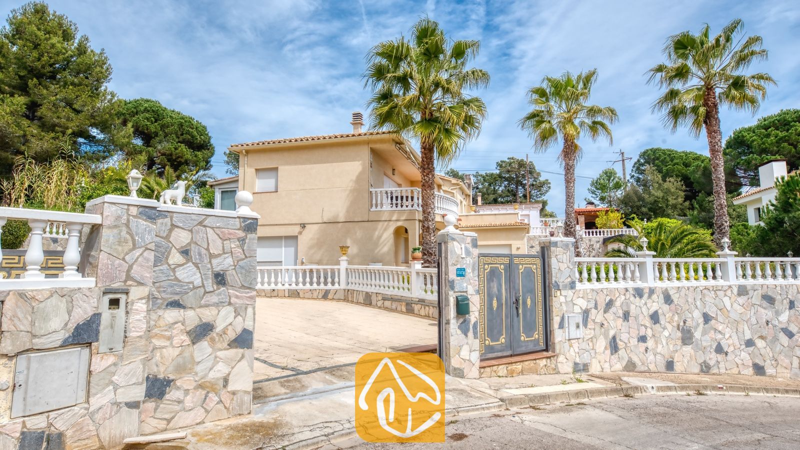 Vakantievilla's Costa Brava Spanje - Villa Estrella - Buitenaanzicht van Villa Estrella met een beige gebouw van twee verdiepingen, een met keien aaneengesloten oprijlaan, decoratieve metalen hekken en hoge palmbomen onder een blauwe hemel.