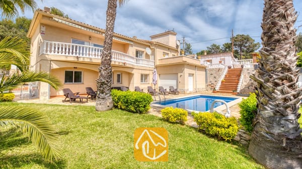 Vakantievilla's Costa Brava Spanje - Villa Estrella - Buitenaanzicht van Villa Estrella met een zwembad, palmbomen, ligstoelen en een beige huis van twee verdiepingen met een balkon.