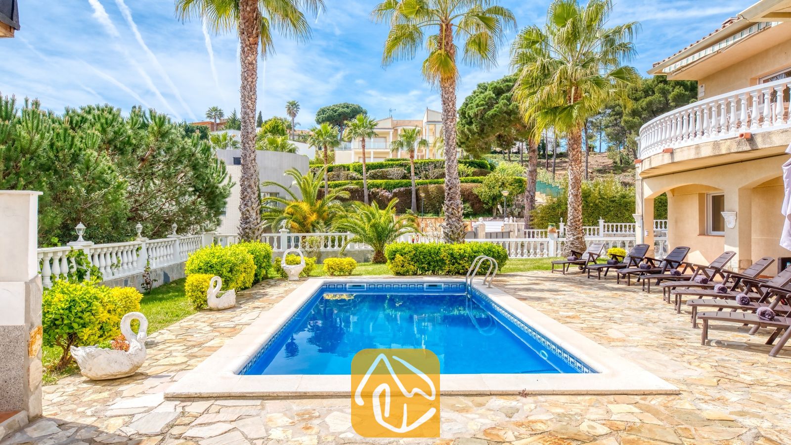 Vakantievilla's Costa Brava Spanje - Villa Estrella - Privézwembad bij Villa Estrella, omringd door een stenen terras, palmbomen, ligstoelen en prachtig onderhouden groene struiken onder een blauwe hemel.