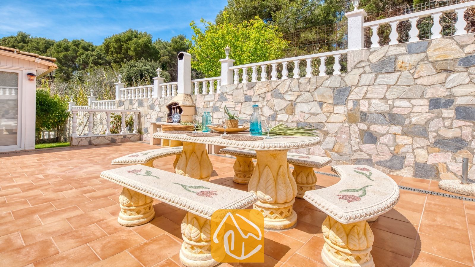 Vakantievilla's Costa Brava Spanje - Villa Estrella - Stenen eettafel en bijpassende mozaïekbanken op een betegeld buitenterras bij Villa Estrella, tegen een achtergrond van een stenen keermuur en bomen.