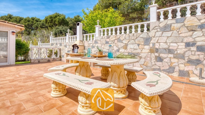 Vakantievilla's Costa Brava Spanje - Villa Estrella - Stenen eettafel en bijpassende mozaïekbanken op een betegeld buitenterras bij Villa Estrella, tegen een achtergrond van een stenen keermuur en bomen.