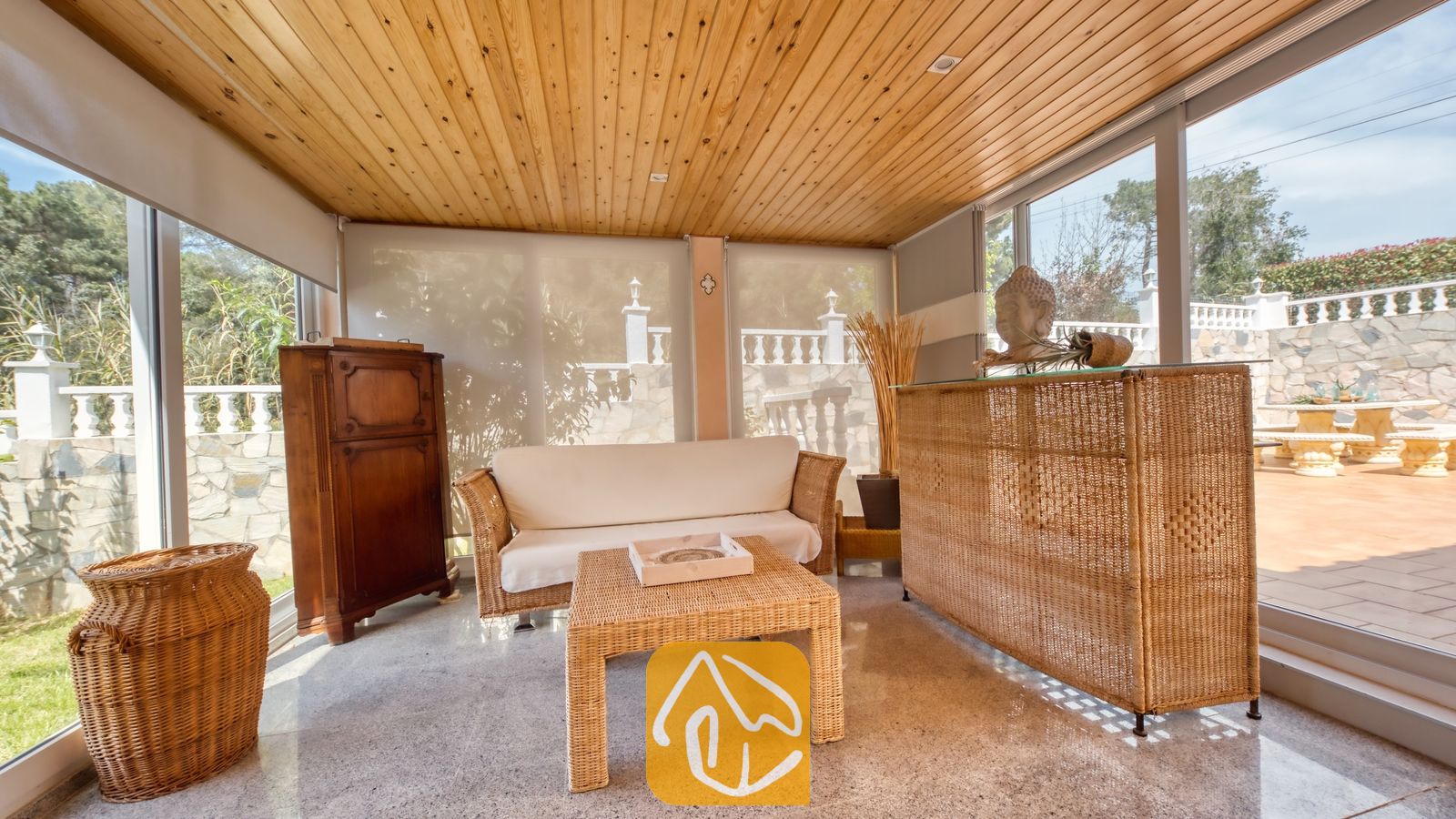 Vakantievilla's Costa Brava Spanje - Villa Estrella - Serre van Villa Estrella met een houten schuin plafond, een rieten bank, een bijpassende salontafel en grote glazen ramen met uitzicht op een stenen terras.