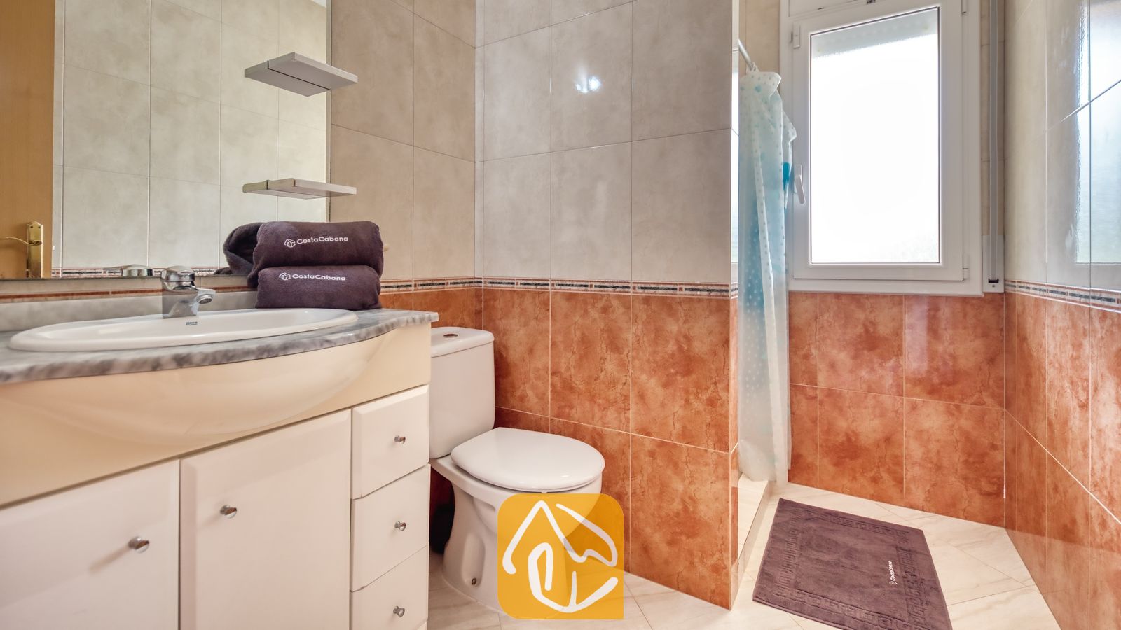 Vakantievilla's Costa Brava Spanje - Villa Estrella - Badkamer voorzien van een wit badkamermeubel met wastafel, toilet en een doucheruimte met een blauw geprint gordijn, betegeld met beige en roodbruine keramische tegels.