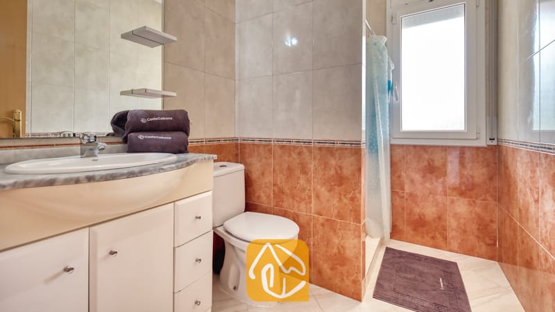 Villas de vacances Costa Brava Espagne - Villa Estrella - Salle de bains comprenant un meuble coiffeur blanc avec lavabo, des toilettes et une douche avec un rideau à motifs bleus, carrelée de céramique beige et brun-rouge.