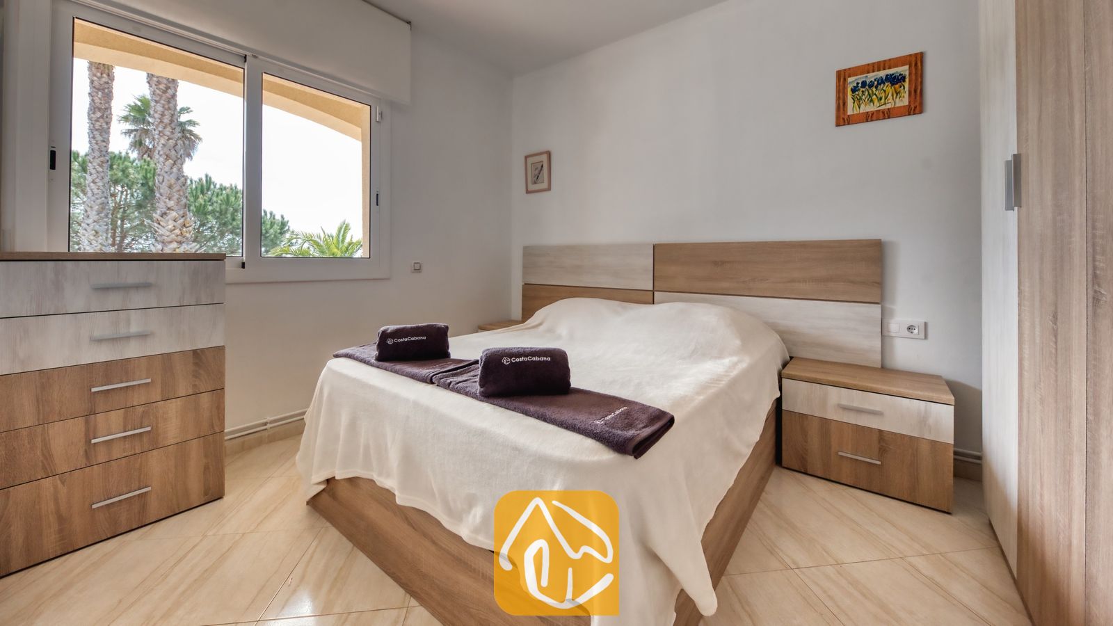 Vakantievilla's Costa Brava Spanje - Villa Estrella - Slaapkamer met een tweepersoonsbed, licht houten meubilair en een raam met uitzicht op palmbomen.