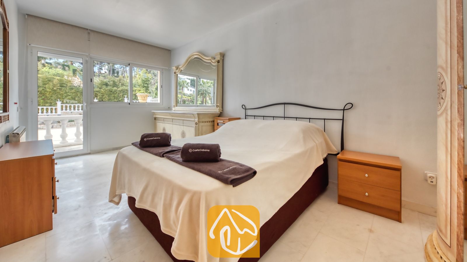 Vakantievilla's Costa Brava Spanje - Villa Estrella - Slaapkamer met een tweepersoonsbed, houten nachtkastjes en grote glazen schuifdeuren die uitkomen op een terras met uitzicht op groene begroeiing.