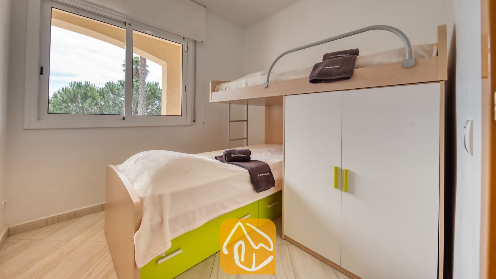 Vakantievilla's Costa Brava Spanje - Villa Estrella - Slaapkamer met een houten stapelbed voorzien van witte en groene opberglades, met opgevouwen donkere handdoeken erop, naast een raam met uitzicht op bomen buiten.