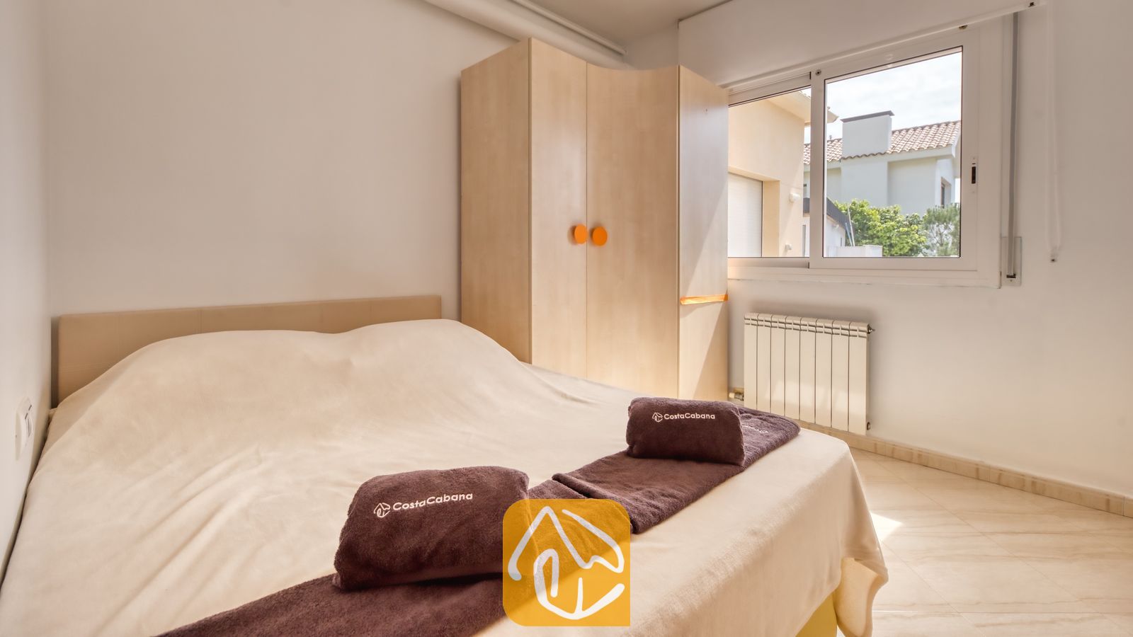 Vakantievilla's Costa Brava Spanje - Villa Estrella - Slaapkamer met een tweepersoonsbed opgemaakt in crèmekleurige lakens, opgevouwen bruine handdoeken met witte tekst, een lichte houten kledingkast met ronde oranje knoppen en een raam.