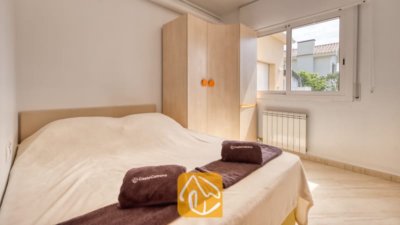 Villas de vacances Costa Brava Espagne - Villa Estrella - Chambre avec un lit double habillé de linge de lit couleur crème, des serviettes marron pliées avec du texte blanc, une armoire en bois clair dotée de poignées rondes orange, et une fenêtre.