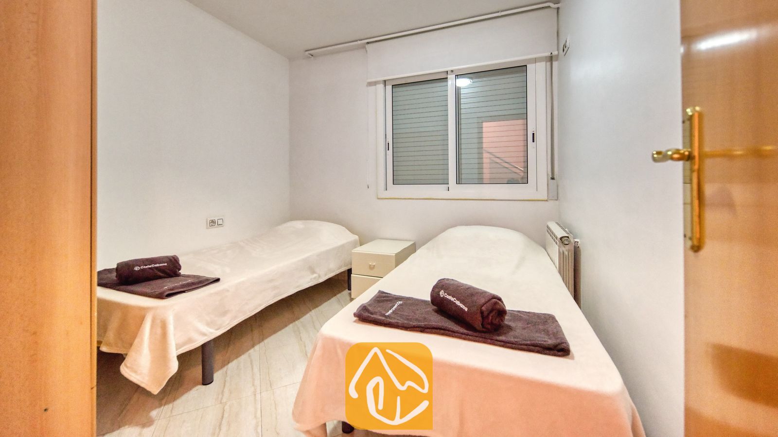 Vakantievilla's Costa Brava Spanje - Villa Estrella - Slaapkamer met twee eenpersoonsbedden opgemaakt met wit linnengoed, een klein wit nachtkastje en een raam met jaloezieën.