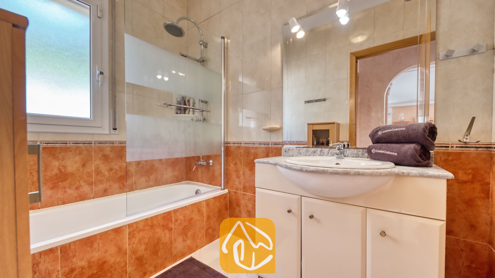 Vakantievilla's Costa Brava Spanje - Villa Estrella - Badkamer voorzien van een wit badkamermeubel met een marmeren blad, een ligbad met een glazen wand en warme roodbruine tegelwanden.