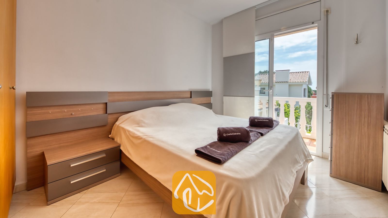 Vakantievilla's Costa Brava Spanje - Villa Estrella - Slaapkamer met een tweepersoonsbed, een houten hoofdbord, geïntegreerde nachtkastjes, opgevouwen bruine handdoeken, tegelvloeren en een glazen deur die naar een balkon leidt.