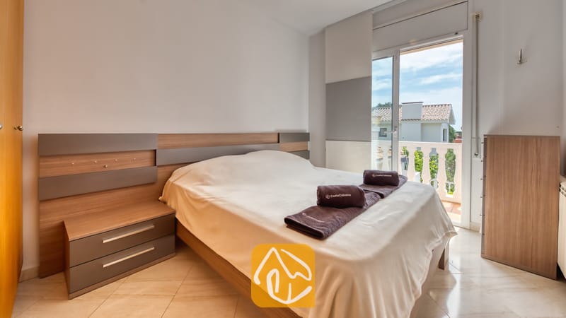 Villas de vacances Costa Brava Espagne - Villa Estrella - Chambre avec un lit double, une tête de lit en bois, des tables de chevet intégrées, des serviettes marron pliées, un sol carrelé et une porte vitrée donnant sur un balcon.