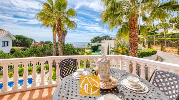 Vakantievilla's Costa Brava Spanje - Villa Estrella - Balkonterras met uitzicht op zee bij Villa Estrella, voorzien van een donker metalen eettafel met een decoratief Boeddha-hoofd als middelpunt, witte tafelsetting en omringende palmbomen.