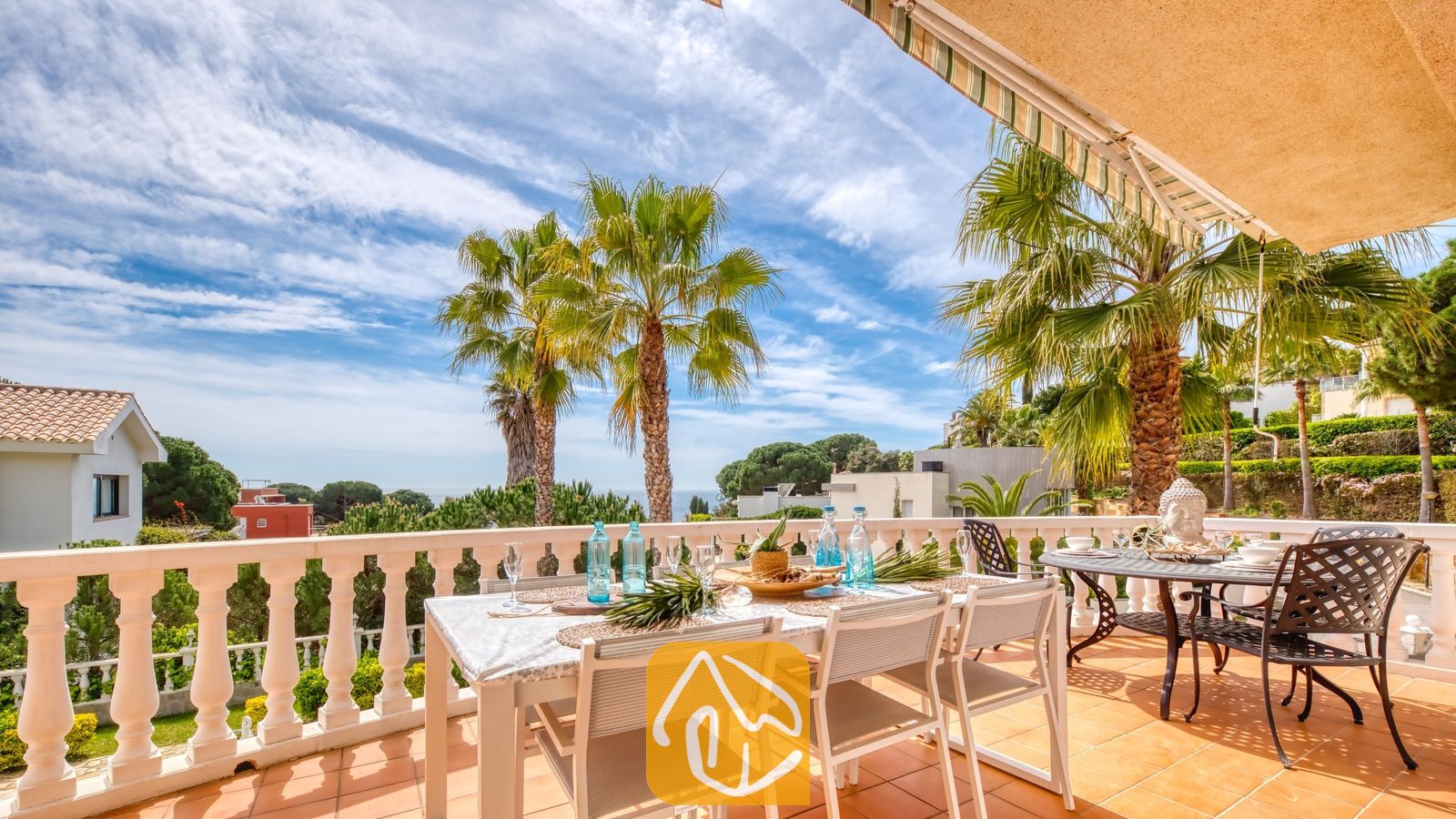 Vakantievilla's Costa Brava Spanje - Villa Estrella - Zonnig terras bij Villa Estrella met een eettafel buiten, gedekt met blauwe glazen flessen, omringd door palmbomen en met uitzicht op zee onder een blauwe hemel.