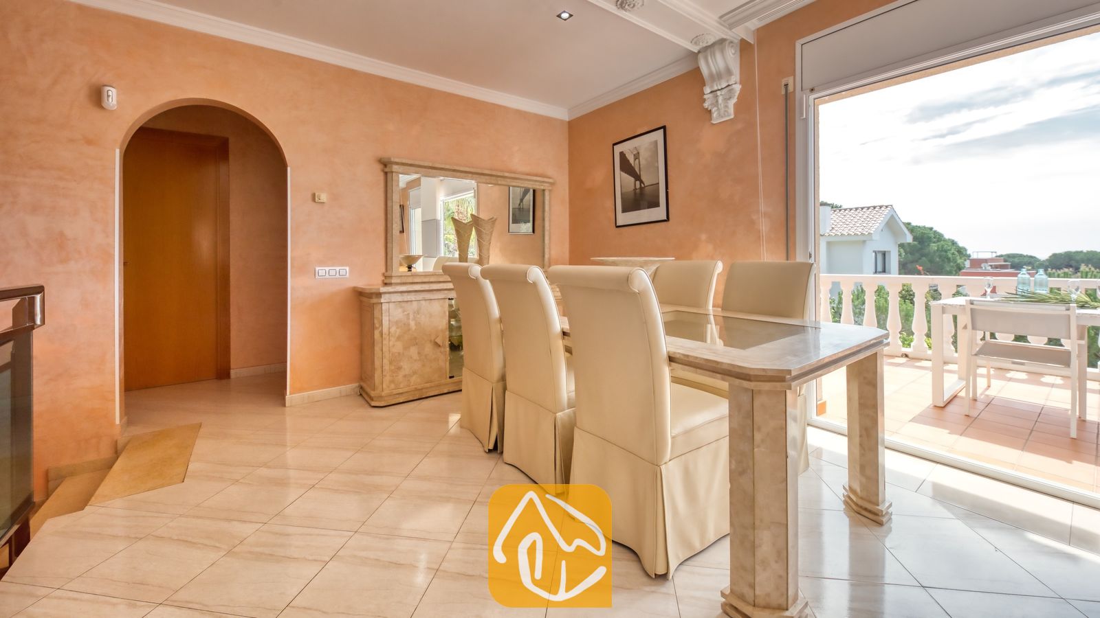 Vakantievilla's Costa Brava Spanje - Villa Estrella - Eethoek in Villa Estrella met een stenen tafel, crème gestoffeerde stoelen, perzikleurige muren en glazen deuren die openstaan naar een balkon.