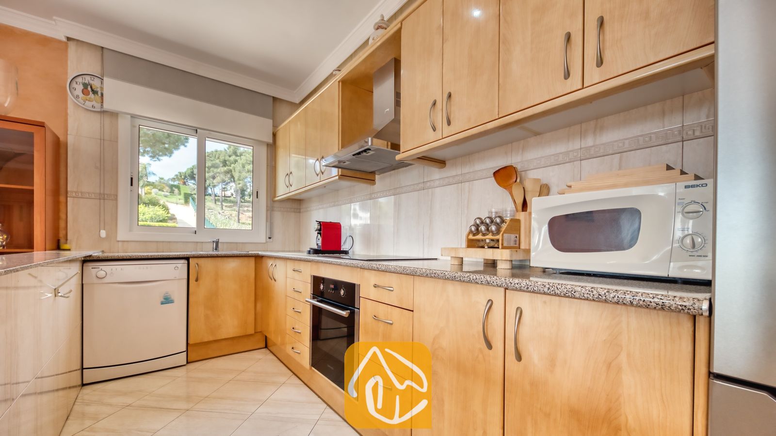 Vakantievilla's Costa Brava Spanje - Villa Estrella - Interieur van de keuken in Villa Estrella met lichthouten kasten, granieten aanrechtbladen, een ingebouwde oven, een witte vaatwasser, een magnetron en een raam met uitzicht op een groen landschap.