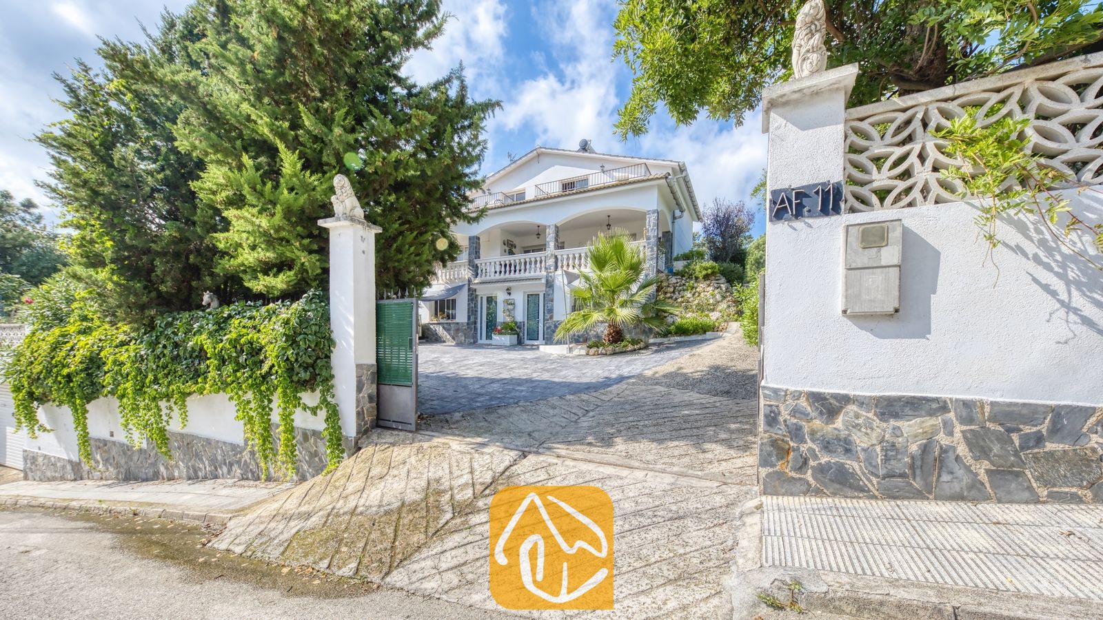 Vakantievilla's Costa Brava Spanje - Villa Geolouk - De oprit aan de buitenzijde en de toegangspoorten van Villa Geolouk, met een wit gebouw van meerdere verdiepingen voorzien van stenen pilaren, balkons en omringd door groene bomen en palmen.