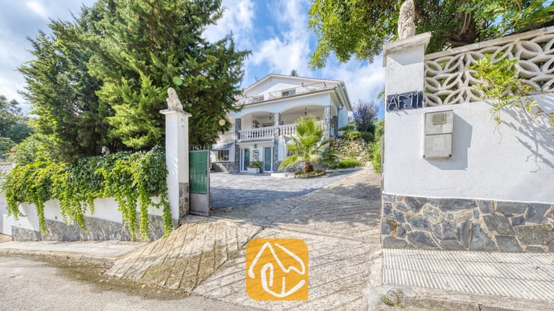Villas de vacances Costa Brava Espagne - Villa Geolouk - Allée extérieure et portail d'entrée de la Villa Geolouk, révélant une maison blanche à plusieurs étages dotée de piliers en pierre et de balcons, nichée au cœur d'une végétation luxuriante composée d'arbres et de palmiers.