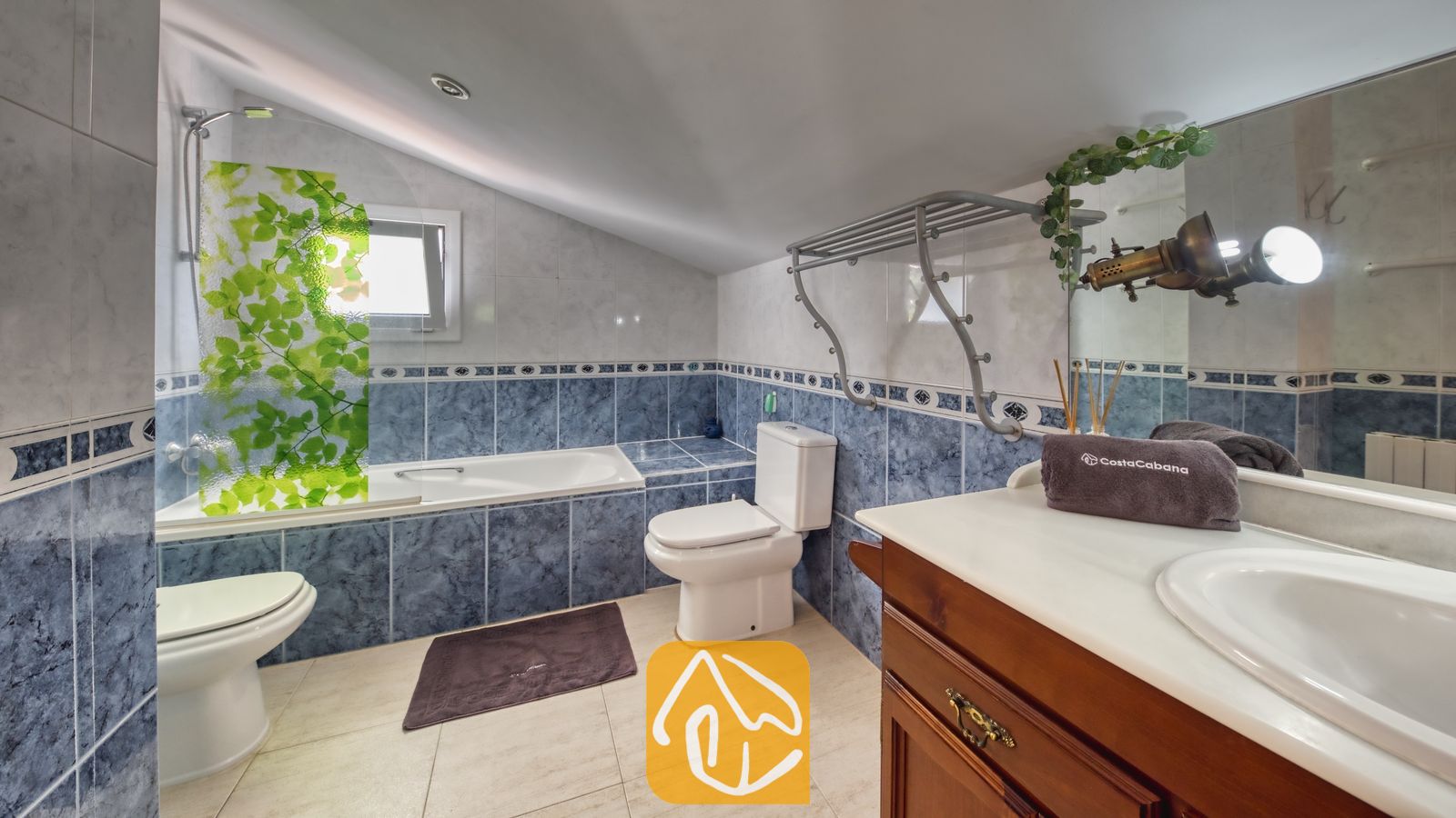 Vakantievilla's Costa Brava Spanje - Villa Geolouk - Badkamer onder het schuine dak met blauwe, marmergetinte tegels, een wit bad met een groen glazen scherm met bladerenmotief, een houten badkamermeubel met een wit aanrechtblad en twee toiletten.