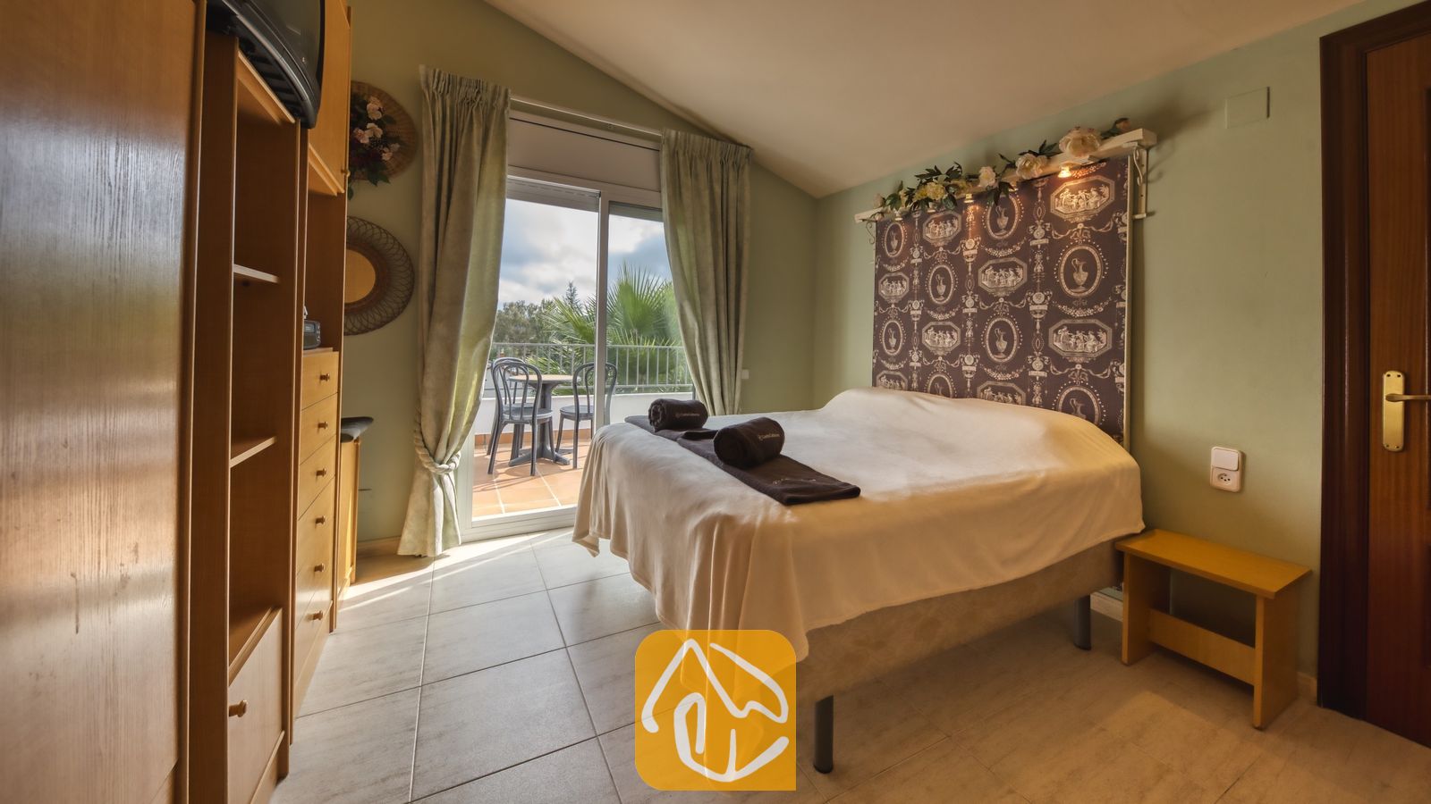 Vakantievilla's Costa Brava Spanje - Villa Geolouk - Slaapkamer met een opgemaakt bed, een gestoffeerd hoofdbord met patroon, tegelvloer en een schuifpui die toegang biedt tot een balkon met een tuintafel en stoelen.