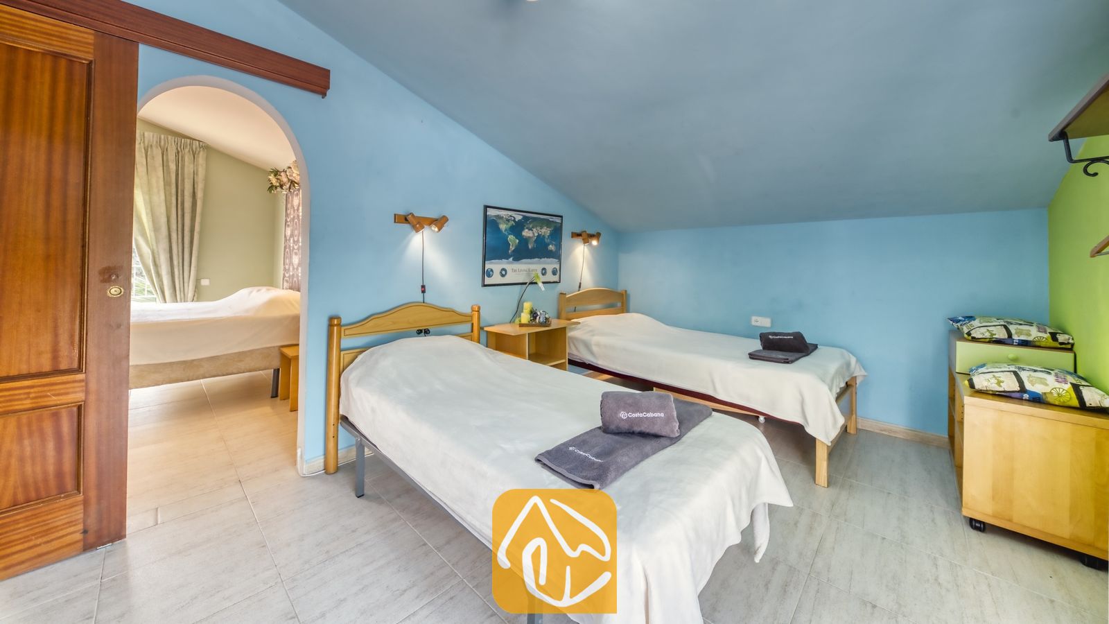 Vakantievilla's Costa Brava Spanje - Villa Geolouk - Slaapkamer met twee houten eenpersoonsbedden, blauwe muren, opgevouwen grijze handdoeken en een toogvormige deuropening die leidt naar een aangrenzende kamer.