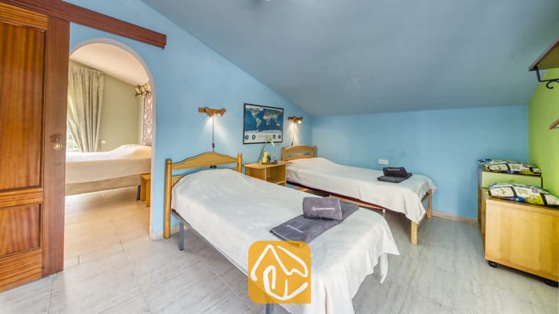 Villas de vacances Costa Brava Espagne - Villa Geolouk - Chambre à coucher dotée de deux lits simples en bois, aux murs bleus, avec des serviettes grises pliées et une porte en arc menant à une pièce attenante.
