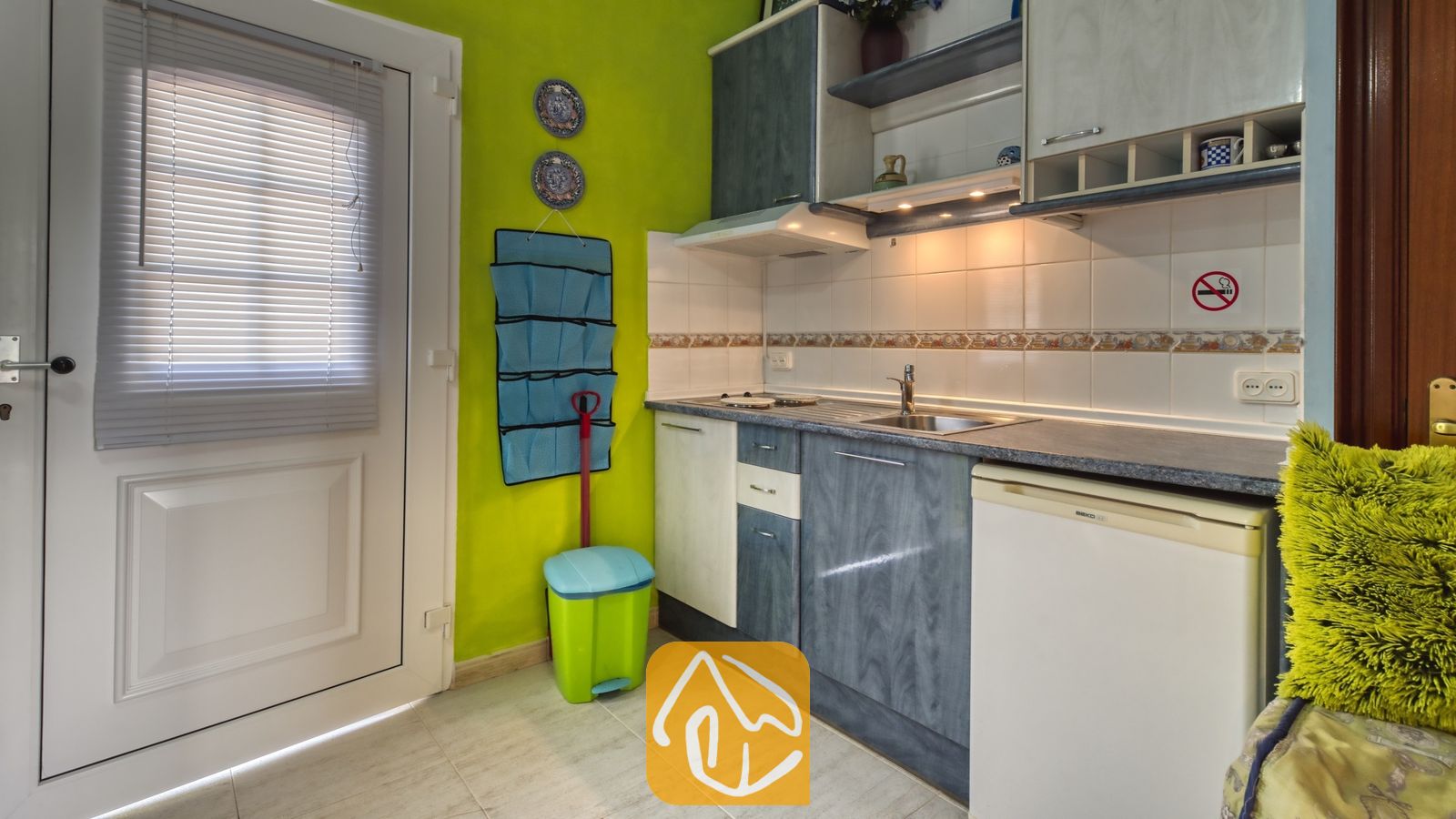 Vakantievilla's Costa Brava Spanje - Villa Geolouk - Compacte keukenhoek met blauw-witte kasten, een roestvrijstalen spoelbak, een tegelachterwand en limoengroene muren naast een witte deur met jaloezieën.