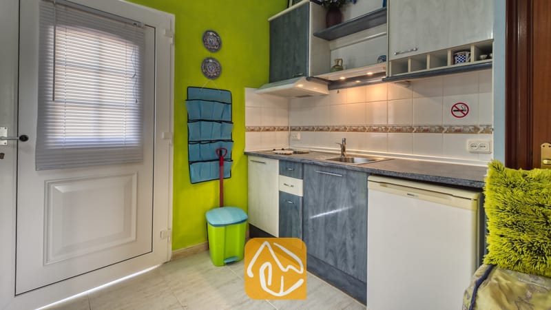 Villas de vacances Costa Brava Espagne - Villa Geolouk - Coin cuisine compact doté de placards bleus et blancs, d'un évier en acier inoxydable, d'une crédence carrelée et de murs vert lime, situé à côté d'une porte blanche avec des stores.