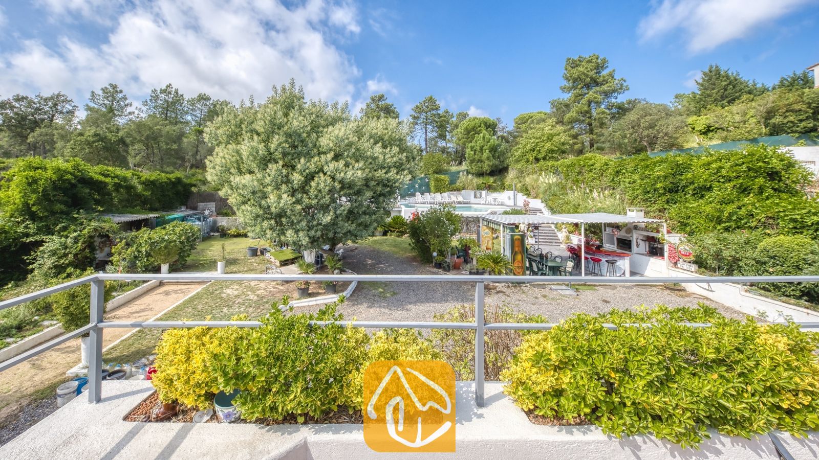 Vakantievilla's Costa Brava Spanje - Villa Geolouk - Overzicht van de weelderige tuin en de buitenbar bij Villa Geolouk, gezien vanaf een balkon met groene potplanten tegen een achtergrond van bomen en een blauwe lucht.