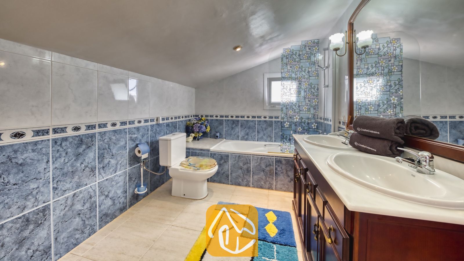 Vakantievilla's Costa Brava Spanje - Villa Geolouk - Badkamer met twee witte wastafels op een donkerhouten badkamermeubel, blauwe marmerlook wandtegels, een ligbad en een toilet onder een schuin plafond.