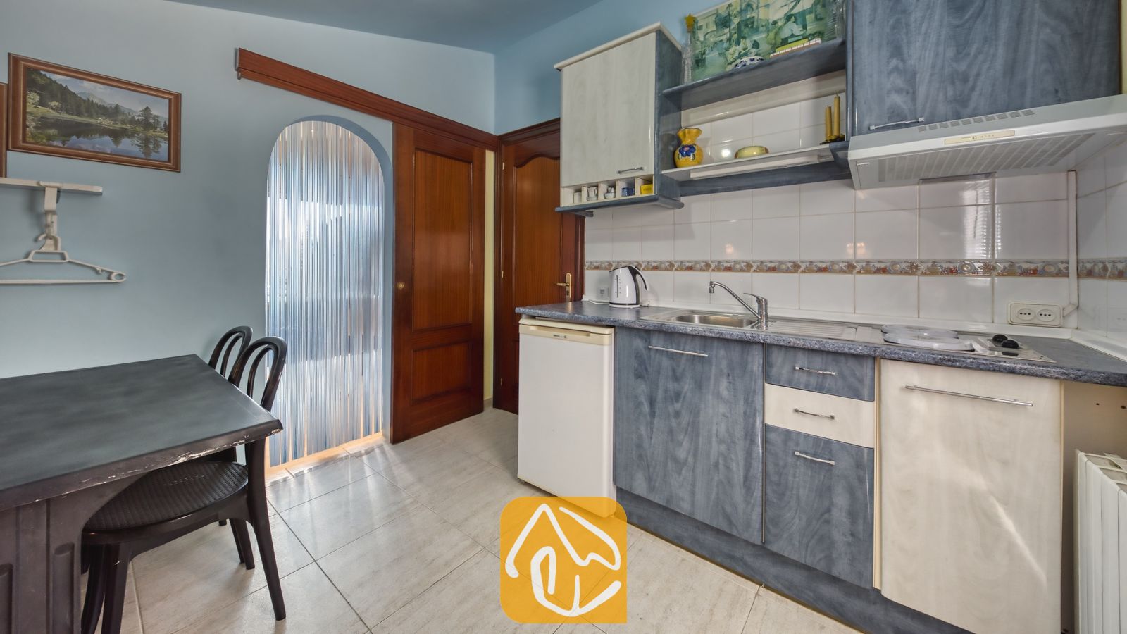 Vakantievilla's Costa Brava Spanje - Villa Geolouk - Compacte keukenhoek met blauw-witte kasten, een eettafel met stoelen, tegelvloeren en een deuropening met een kralengordijn.