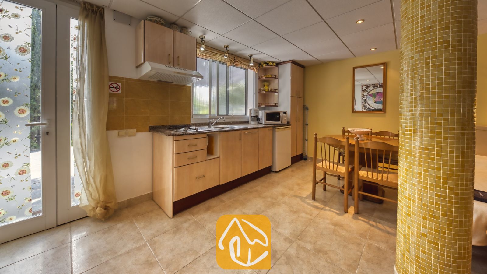 Vakantievilla's Costa Brava Spanje - Villa Geolouk - Keuken en eethoek met lichte houten kasten, tegelvloeren, een gele mozaïekzuil en een houten tafel met bijpassende stoelen.